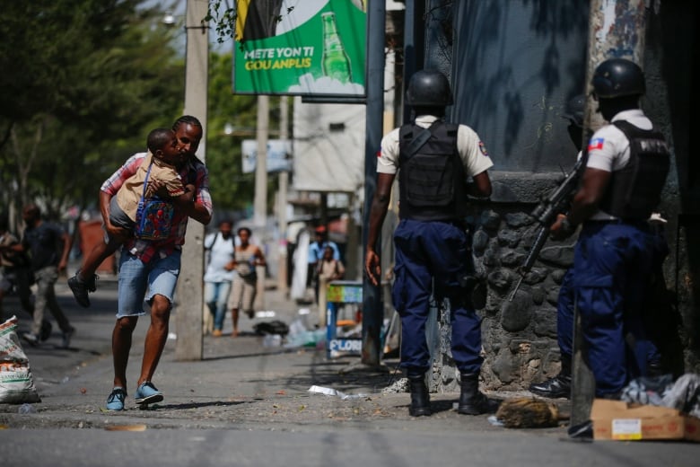 A parent, carrying his child after picking him up from school, runs past police as they carry out an operation against gangs in the Bel-Air area of Port-au-Prince, Haiti, Friday, March 3, 2023. (AP Photo/Odelyn Joseph)